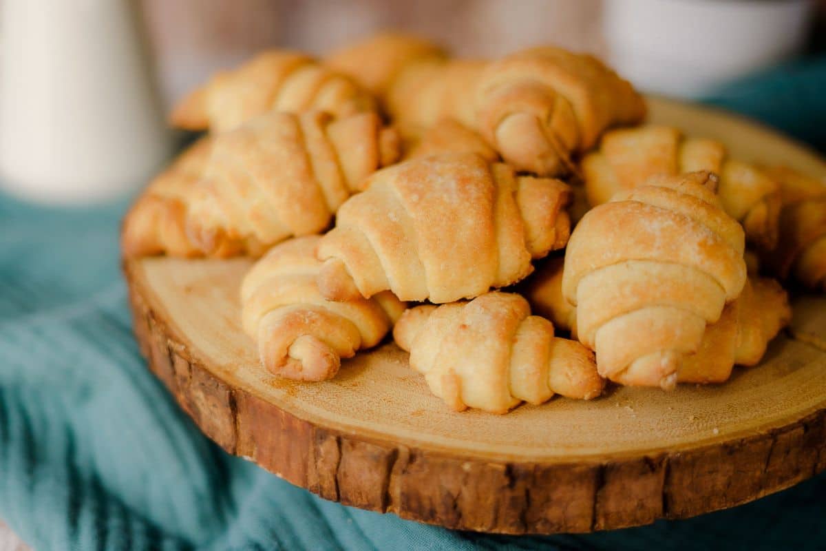 A wooden platter holds a stack of golden-brown croissants on a teal cloth, perfect for adding a touch of elegance to your Thanksgiving table.