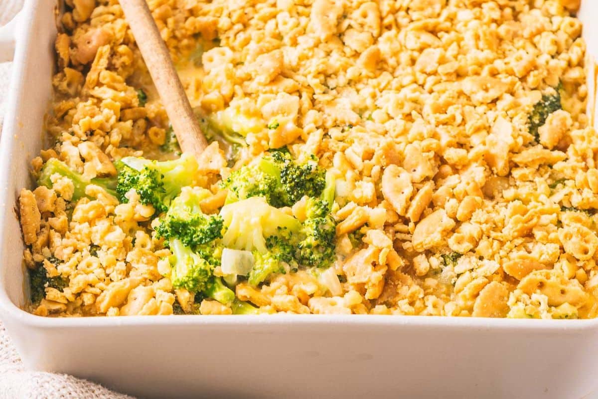 Gluten-free broccoli casserole topped with crumbled crackers in a baking dish, perfect for Thanksgiving. A wooden spoon rests in the corner, ready for serving.