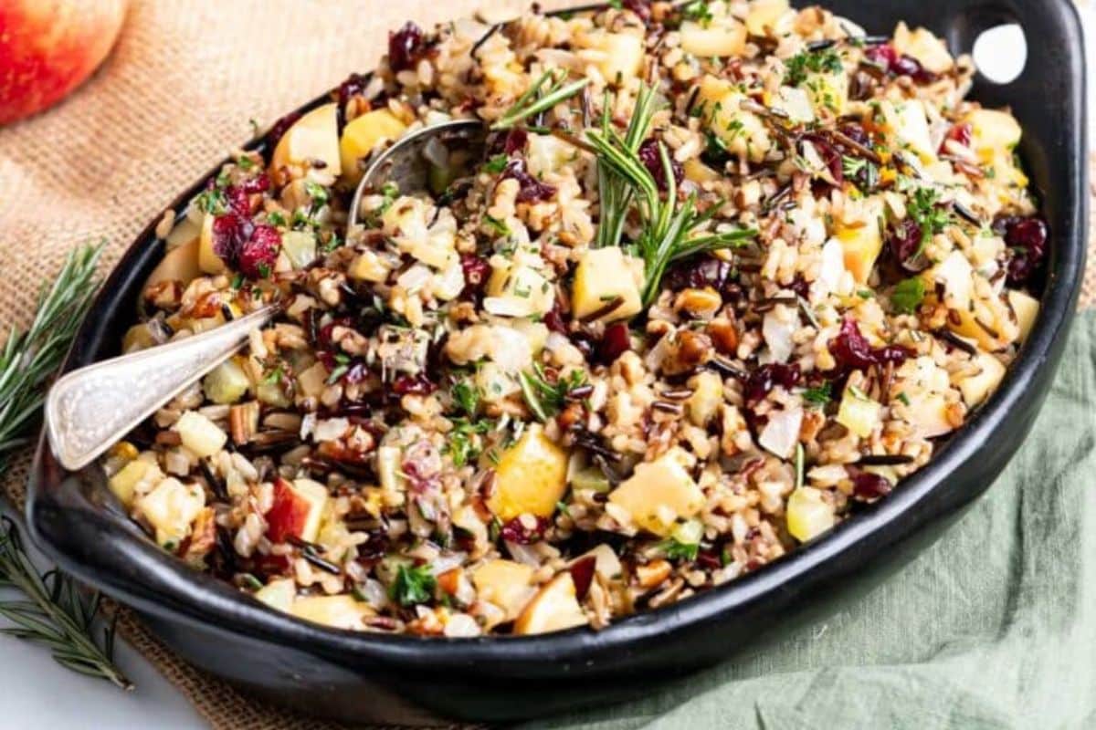 A bowl of wild rice salad with apples, cranberries, nuts, and herbs sits invitingly on a black dish, gluten-free and perfect for Thanksgiving.
