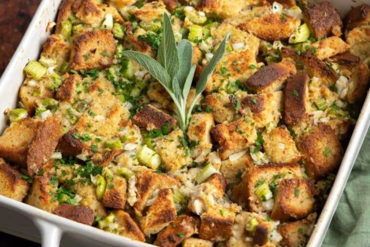 Thanksgiving baked stuffing with browned bread cubes, chopped celery, and herbs, garnished with a sprig of fresh sage in a white casserole dish. Perfect for those seeking delicious Gluten-Free recipes to celebrate the holiday.