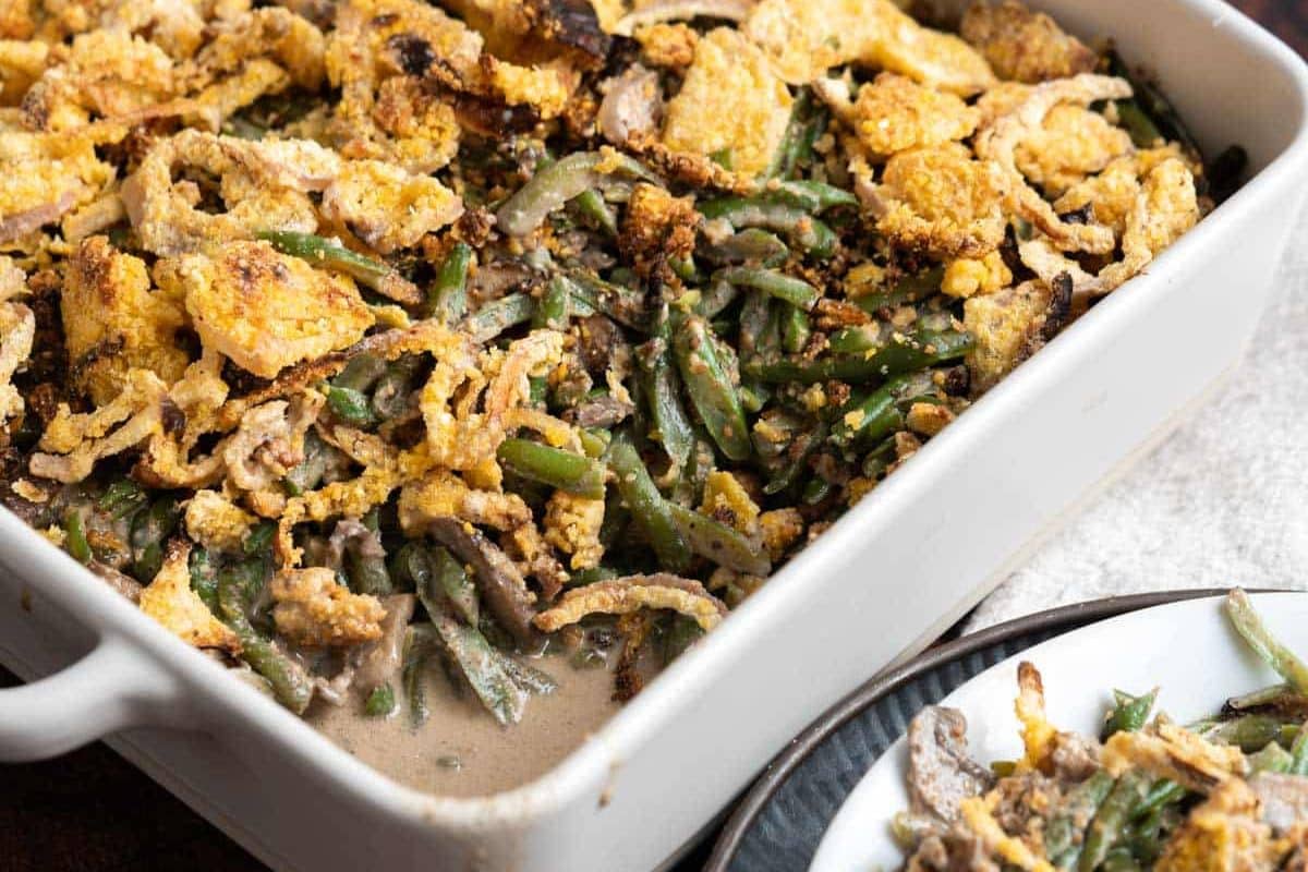 A casserole dish brimming with green bean casserole topped with crispy fried onions sits beside a plate showcasing a tempting serving, perfect for Thanksgiving gatherings. This gluten-free recipe is sure to be a holiday favorite!