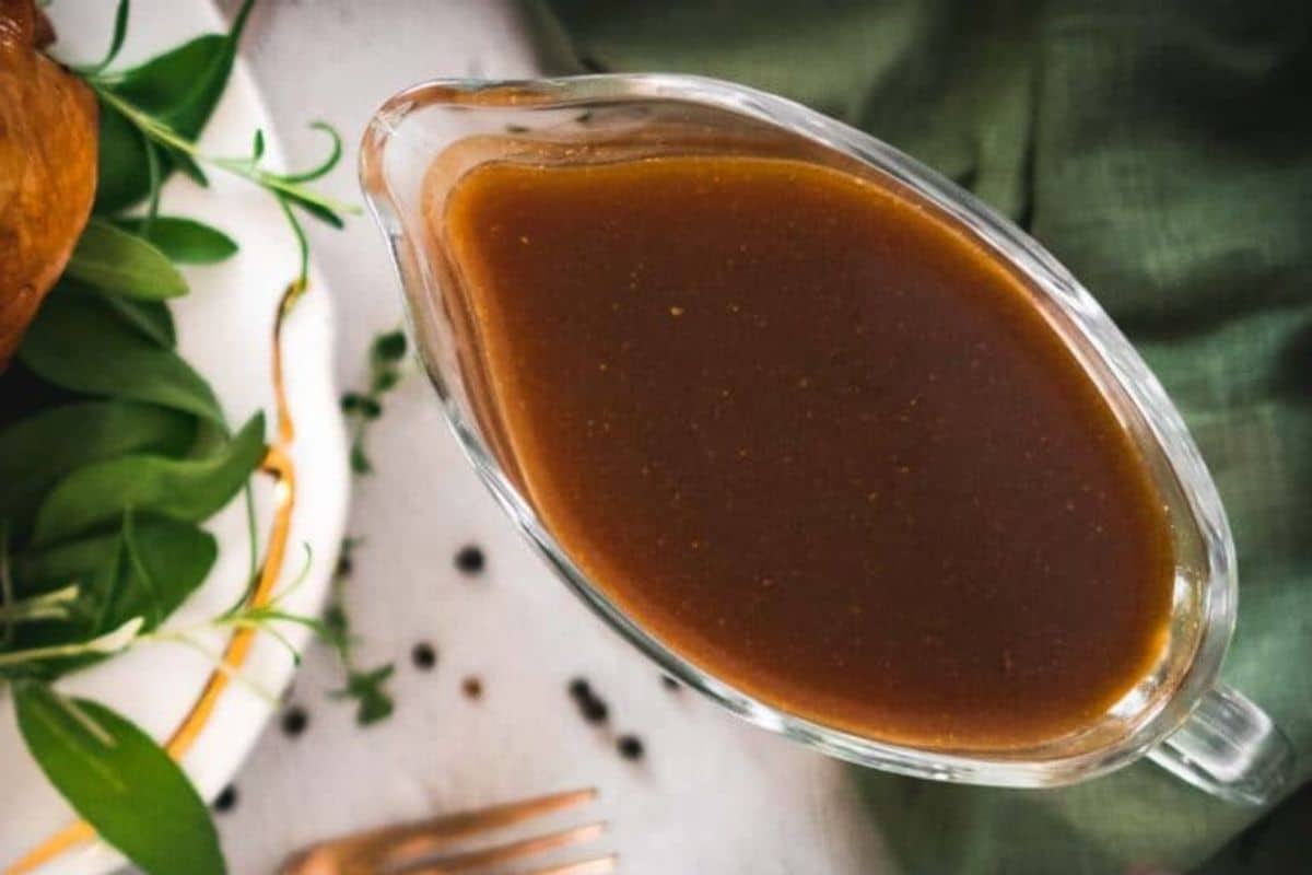 A glass gravy boat filled with rich, brown gravy sits beside a plate adorned with green leaves and a fork on a festive tablecloth, perfect for serving up your favorite gluten-free recipes this Thanksgiving.