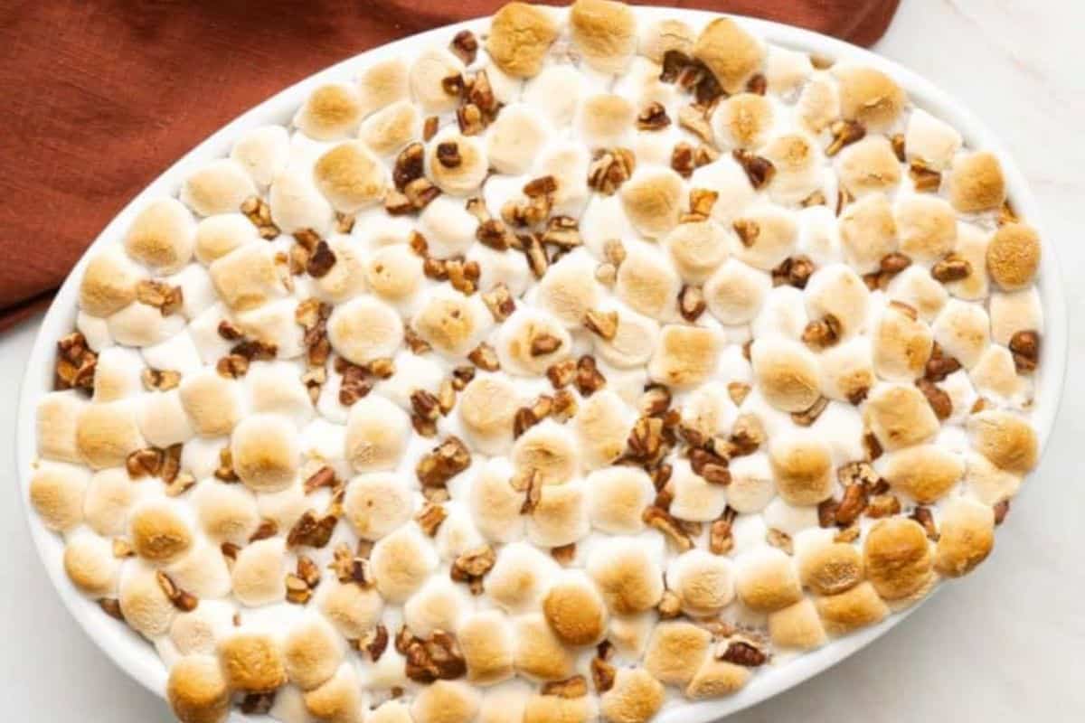 A gluten-free casserole dish filled with toasted marshmallows and pecans on top of a creamy base, perfect for Thanksgiving, set on a white surface with a brown cloth in the background.
