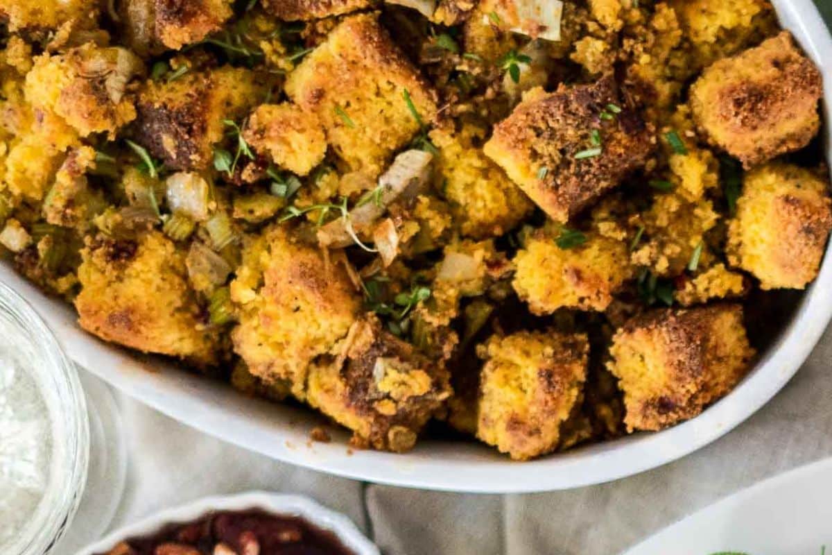 A bowl of cornbread stuffing with herbs and diced onions sits on a table, surrounded by other Thanksgiving side dishes.