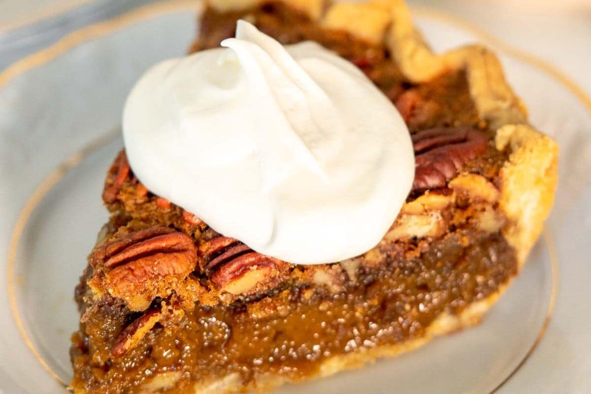 A slice of pecan pie, perfect for Thanksgiving, is topped with a swirl of whipped cream on a white plate.