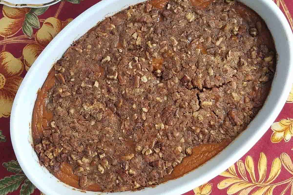 Oval dish of sweet potato casserole topped with a crumbly pecan and brown sugar mixture, perfect for Thanksgiving, on a red tablecloth with fruit patterns. This gluten-free delight will be the star of your holiday table.