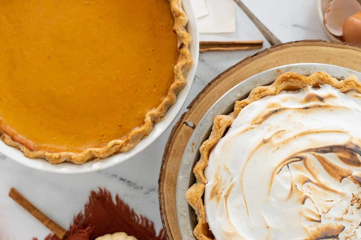 Two pies on a table: pumpkin pie on the left and meringue-topped pie on the right, perfect for a Thanksgiving feast. Nearby are cinnamon sticks and a brown cloth, adding to the cozy atmosphere with gluten-free recipe options in mind.
