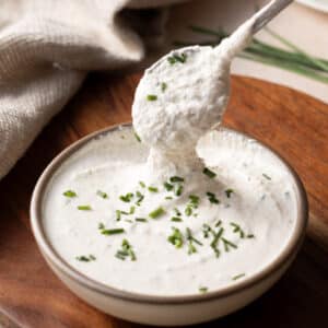 A spoon scoops creamy horseradish sauce from a bowl, garnished with chopped chives, on a wooden surface.