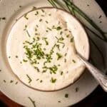 A bowl of creamy horseradish mustard sauce garnished with chopped chives sits on a speckled plate, accompanied by fresh chive stalks. A silver spoon rests elegantly on the bowl's edge.
