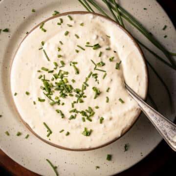 A bowl of creamy horseradish mustard sauce garnished with chopped chives sits on a speckled plate, accompanied by fresh chive stalks. A silver spoon rests elegantly on the bowl's edge.