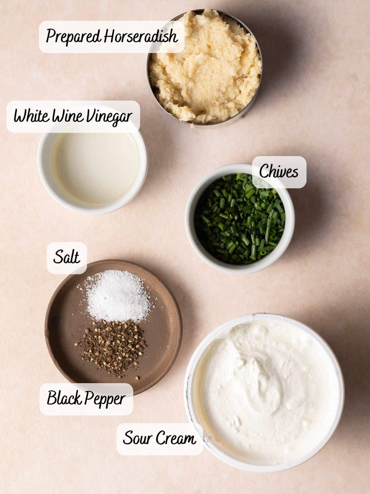 Ingredients for a zesty horseradish sauce, featuring prepared horseradish, white wine vinegar, chives, sour cream, salt, and black pepper artfully arranged on a light surface.