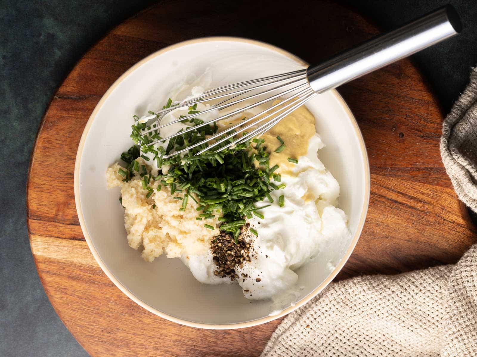 A bowl with sour cream, horseradish mustard sauce, chopped chives, black pepper, and mashed potatoes is surrounded by a whisk on a wooden board with a cloth nearby.