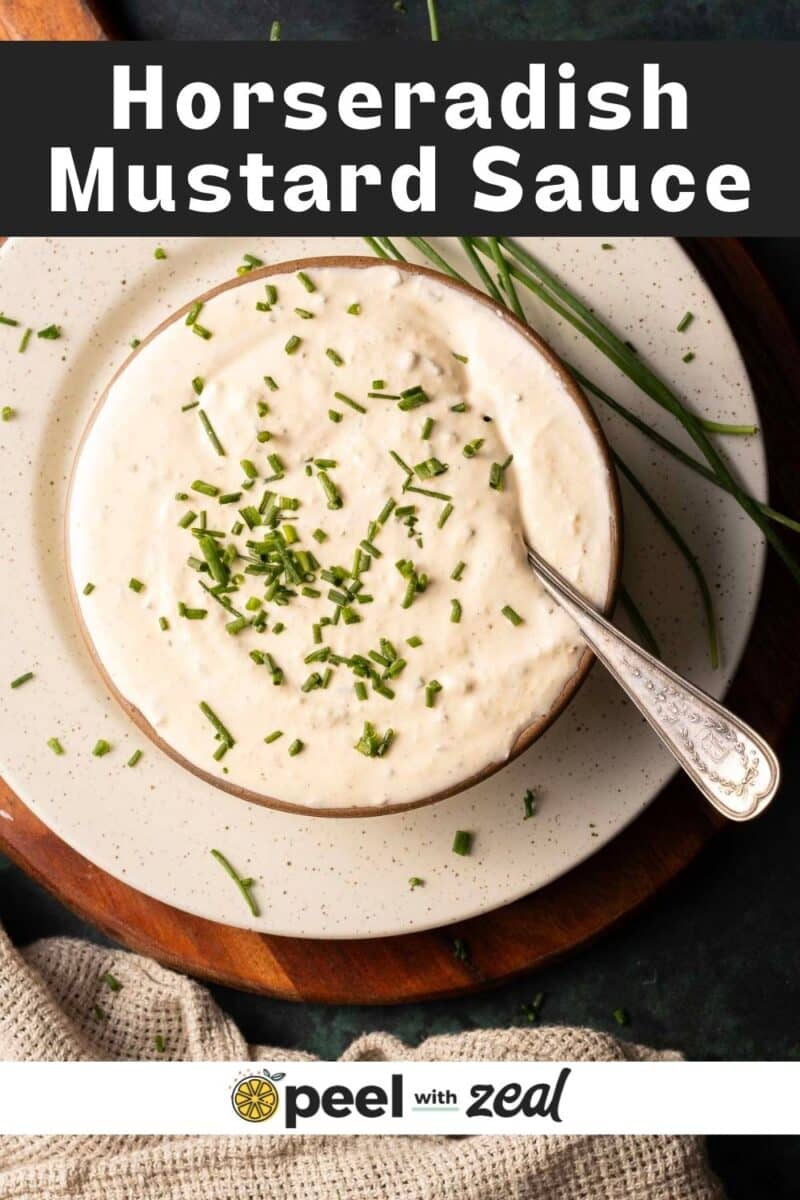 A bowl of zesty horseradish mustard sauce crowned with chopped chives sits elegantly on a plate, accompanied by a spoon ready for dipping.
