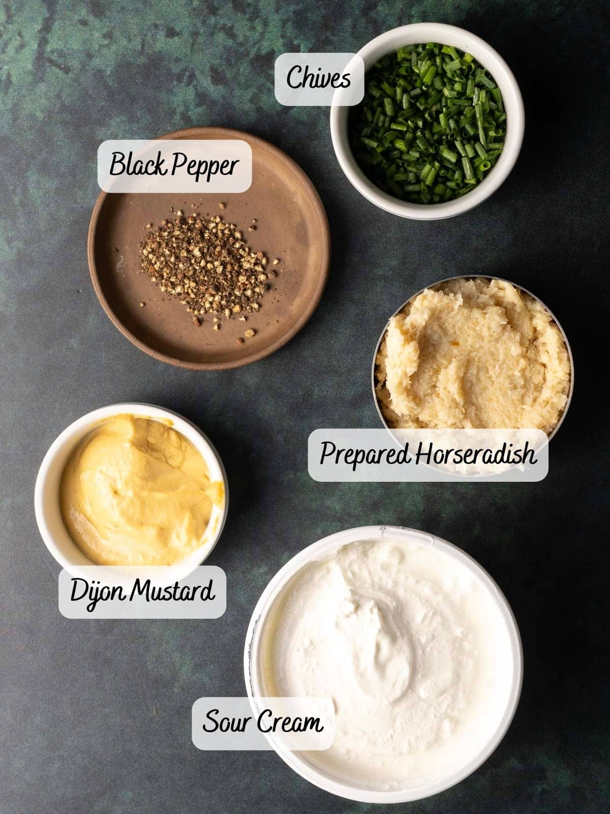 Five ingredients are arranged on a dark surface: chopped chives, a plate of black pepper, prepared horseradish, Dijon mustard, and sour cream—perfect elements for crafting a zesty Horseradish Mustard Sauce.