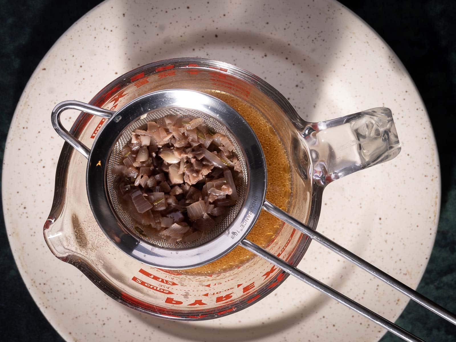 A metal strainer sits on a glass measuring cup filled with rich au jus, filtering chopped cooked onions above a speckled ceramic plate.