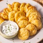 A plate of golden, round mashed potato puffs arranged in a spiral pattern, delicately garnished with rosemary. A small bowl of creamy white dip with chopped herbs and a hint of Parmesan is placed on the side.