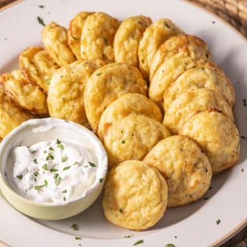 A plate of golden, round mashed potato puffs arranged in a spiral pattern, delicately garnished with rosemary. A small bowl of creamy white dip with chopped herbs and a hint of Parmesan is placed on the side.
