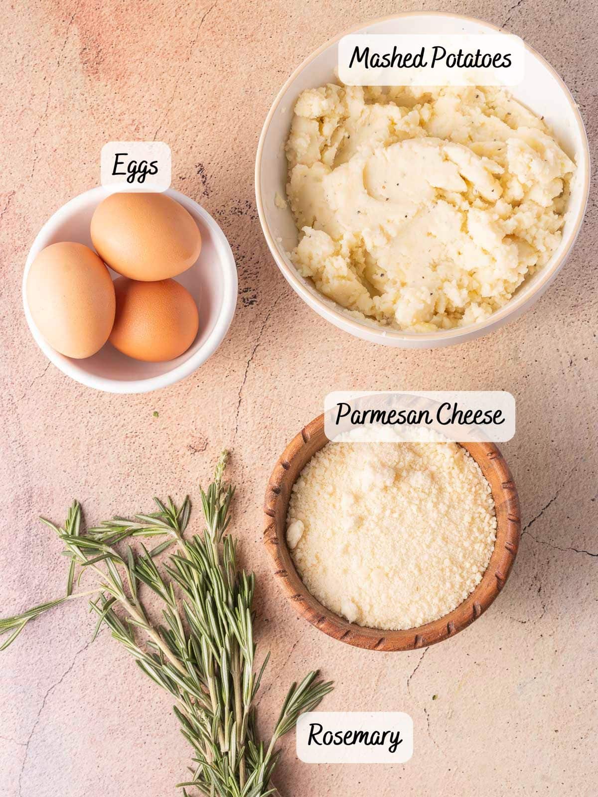 A bowl of eggs, a bowl of mashed potato puffs, a generous sprinkle of Parmesan cheese, and sprigs of rosemary rest gracefully on a textured surface.