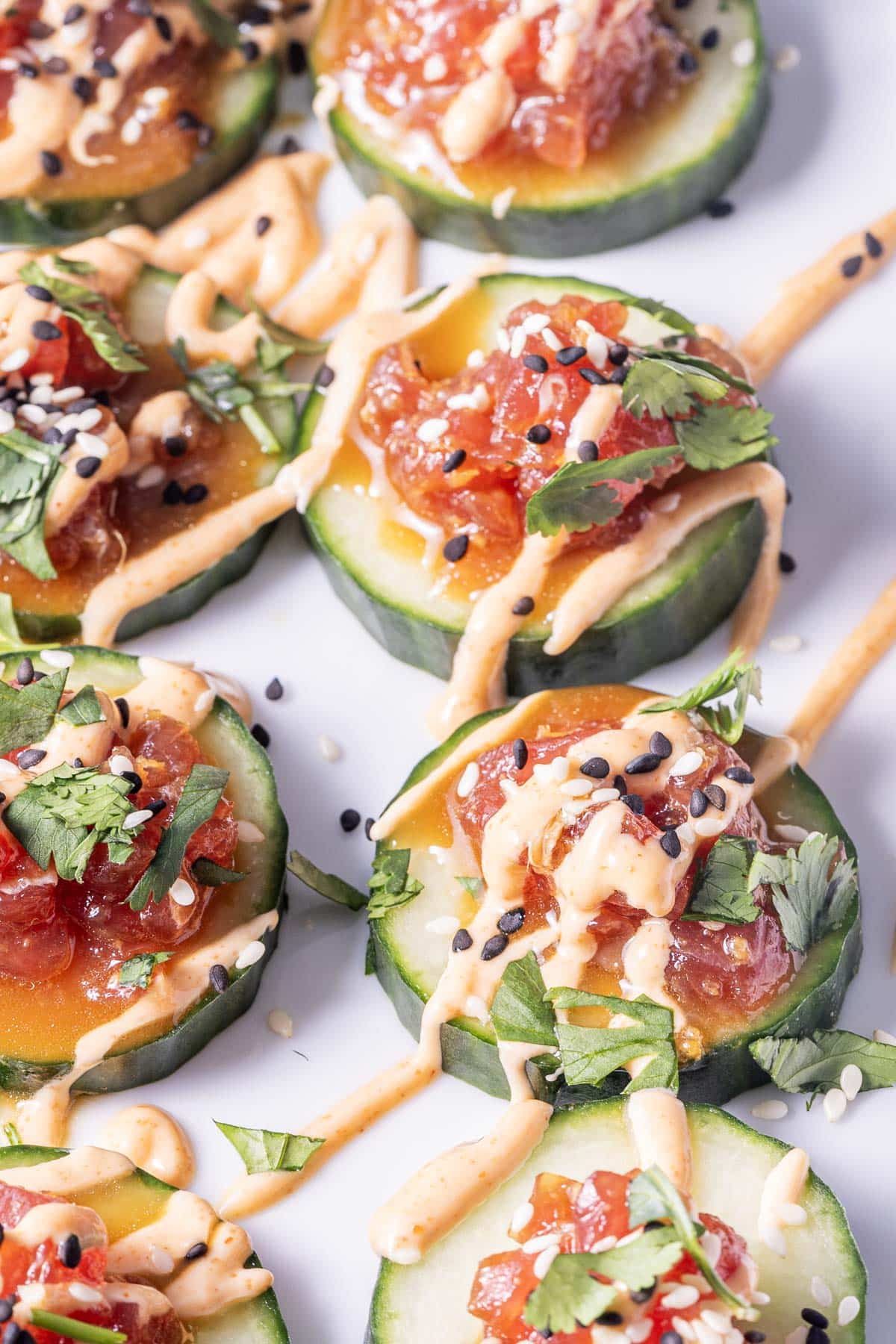 Cucumber slices topped with diced vegetables, drizzled with creamy sauce, and garnished with sesame seeds and cilantro.