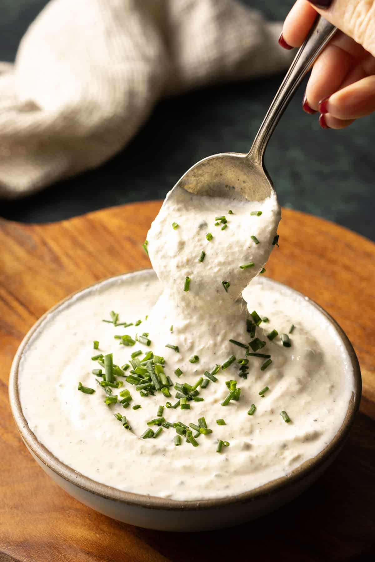 A hand holding a spoon dips into a bowl of creamy horseradish mustard sauce topped with chopped chives on a wooden surface.