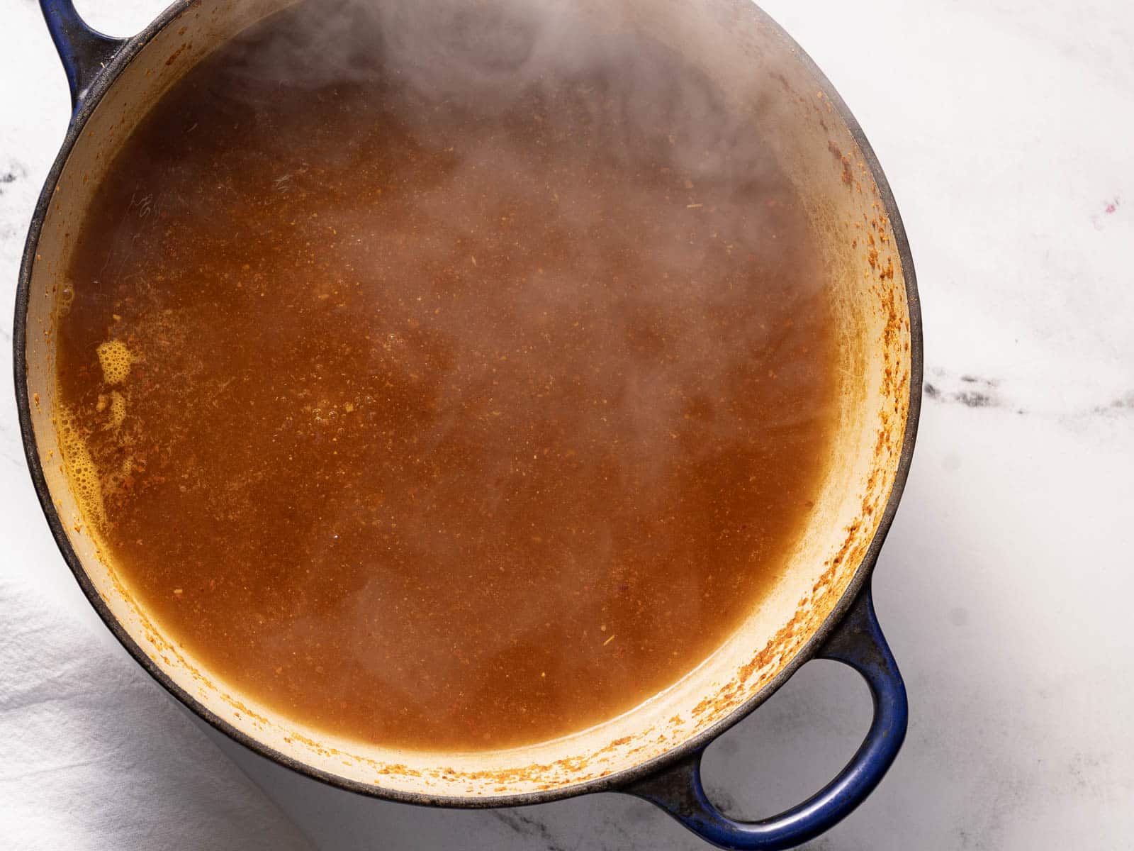 A steaming pot of brown broth on a stovetop with a blue handle, placed on a white surface.