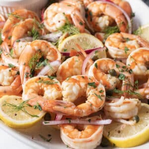 A plate of cooked shrimp garnished with lemon slices, red onion, dill, and capers.
