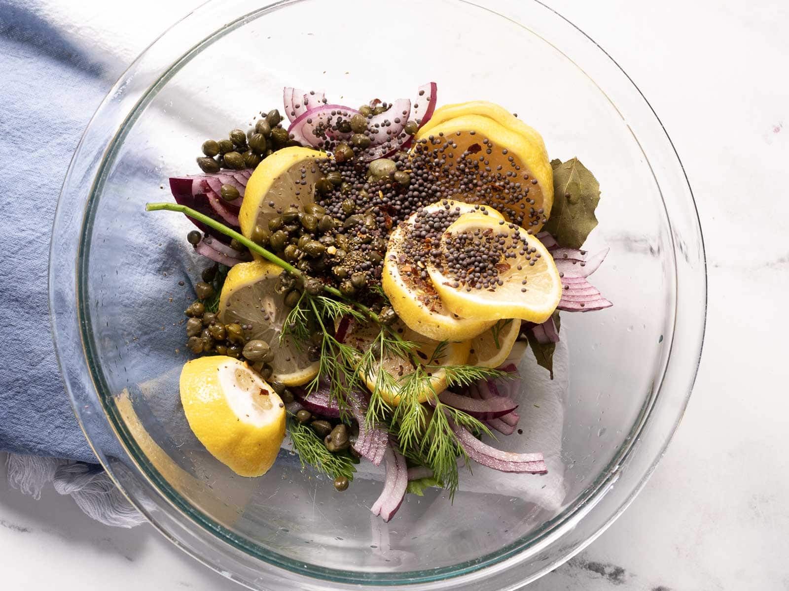 Glass bowl with sliced lemons, capers, mustard seeds, red onion, dill, and bay leaves on a marble surface with a blue cloth.