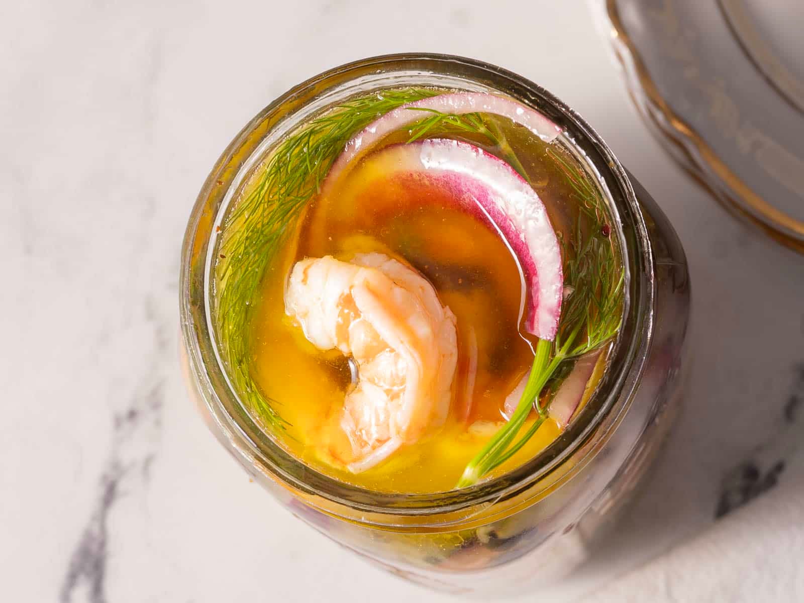 Jar of pickled shrimp with a sprig of dill and sliced red onions in a clear brine, placed on a marble surface next to a plate.