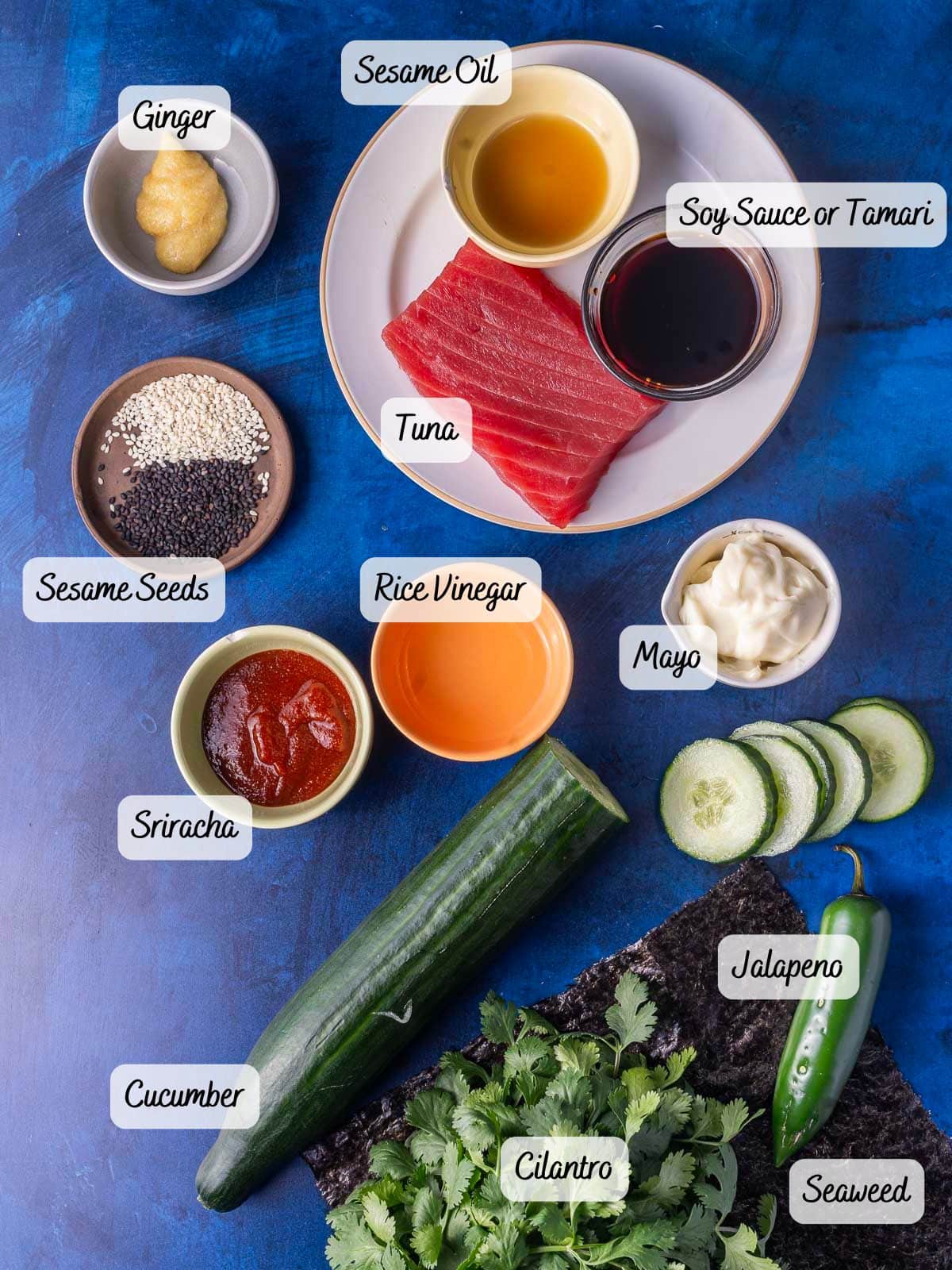 Ingredients for a dish include tuna, soy sauce, sesame oil, rice vinegar, mayo, ginger, sesame seeds, cucumber, jalapeno, cilantro, seaweed, and sriracha on a blue surface.