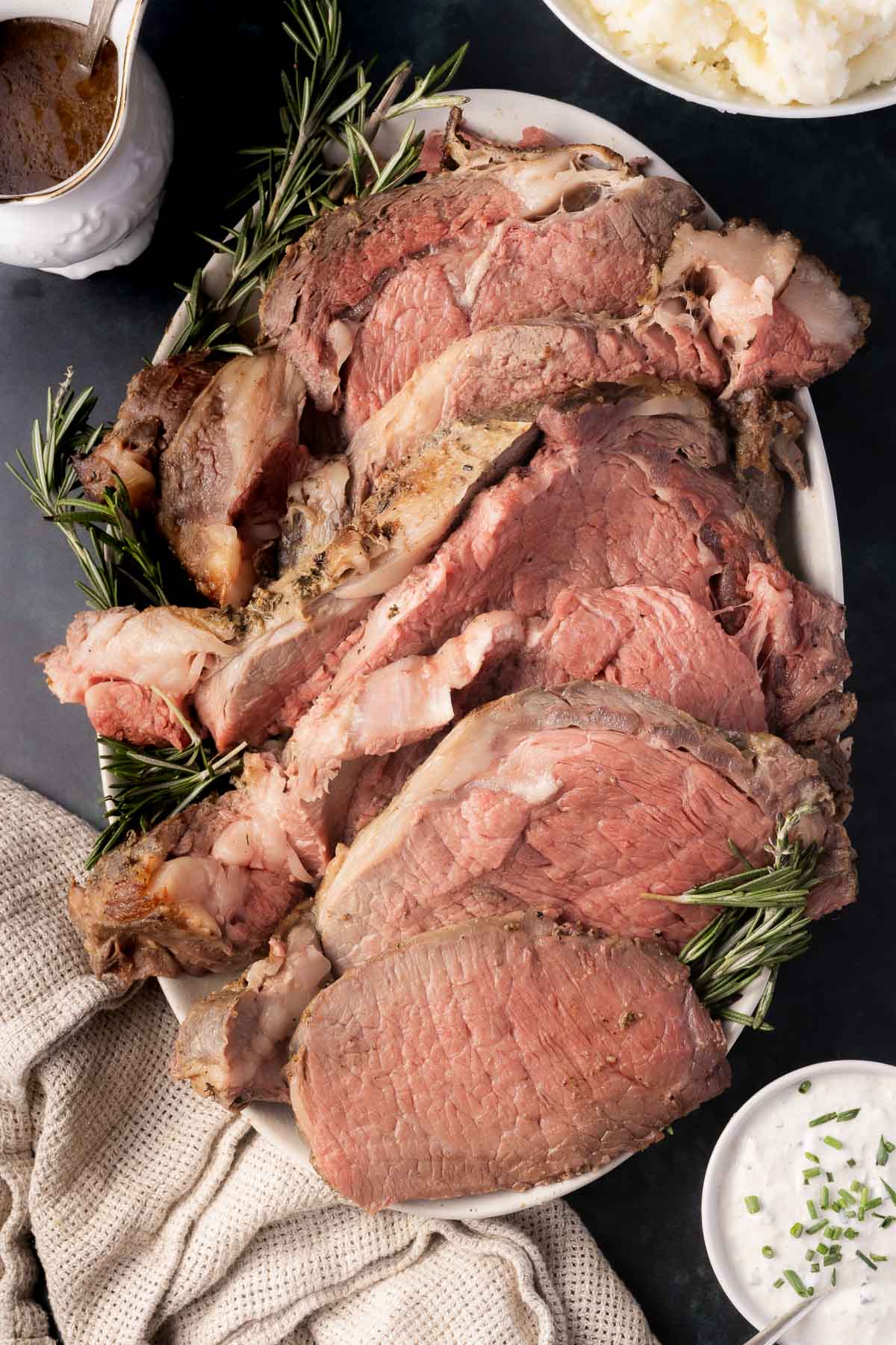 A platter of sous vide prime rib, thinly sliced and garnished with rosemary, accompanied by horseradish sauce and a bowl of mashed potatoes.