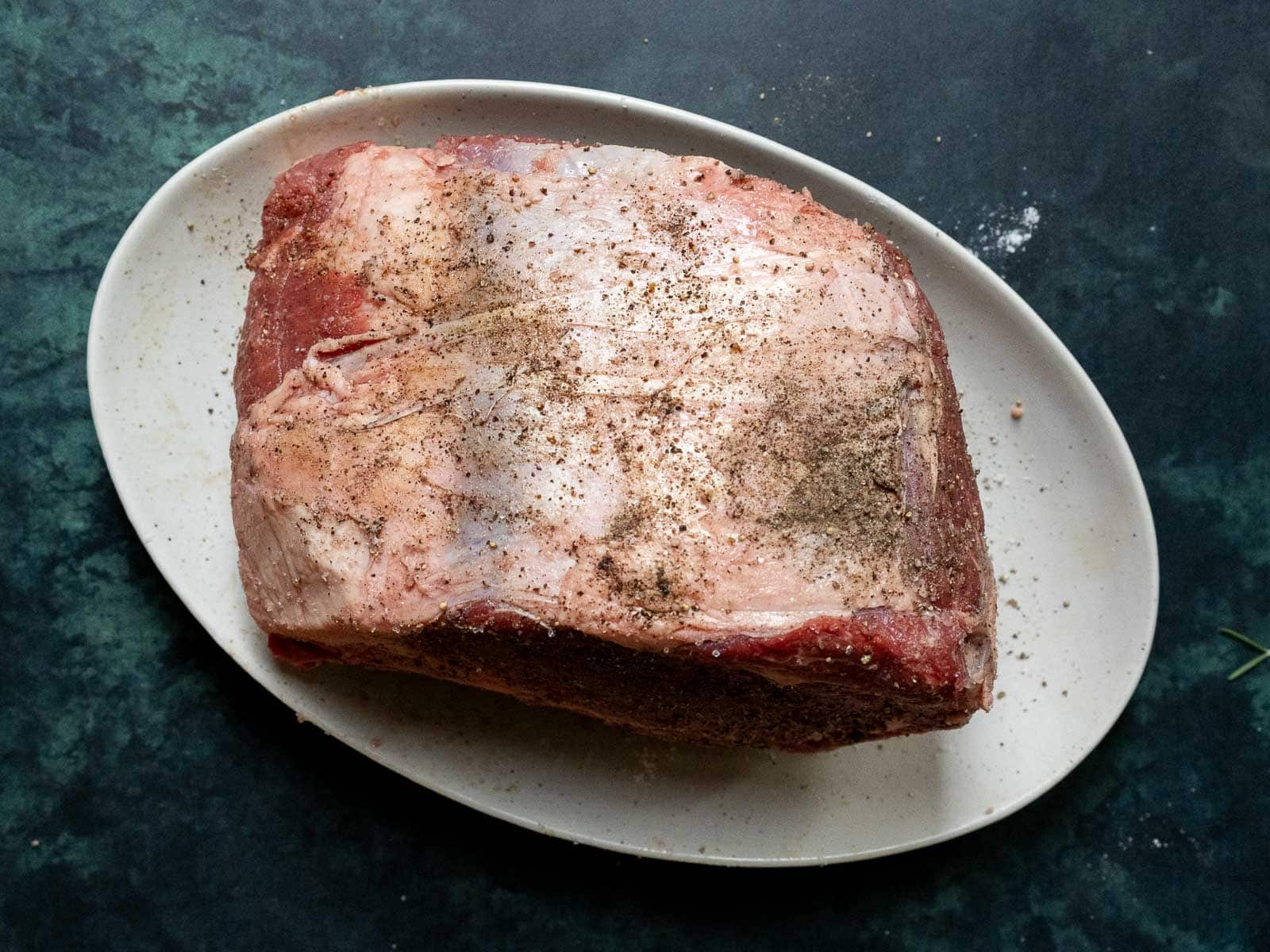 A raw prime rib roast on a white oval plate, seasoned with salt and pepper, ready for the sous vide method on a dark countertop.