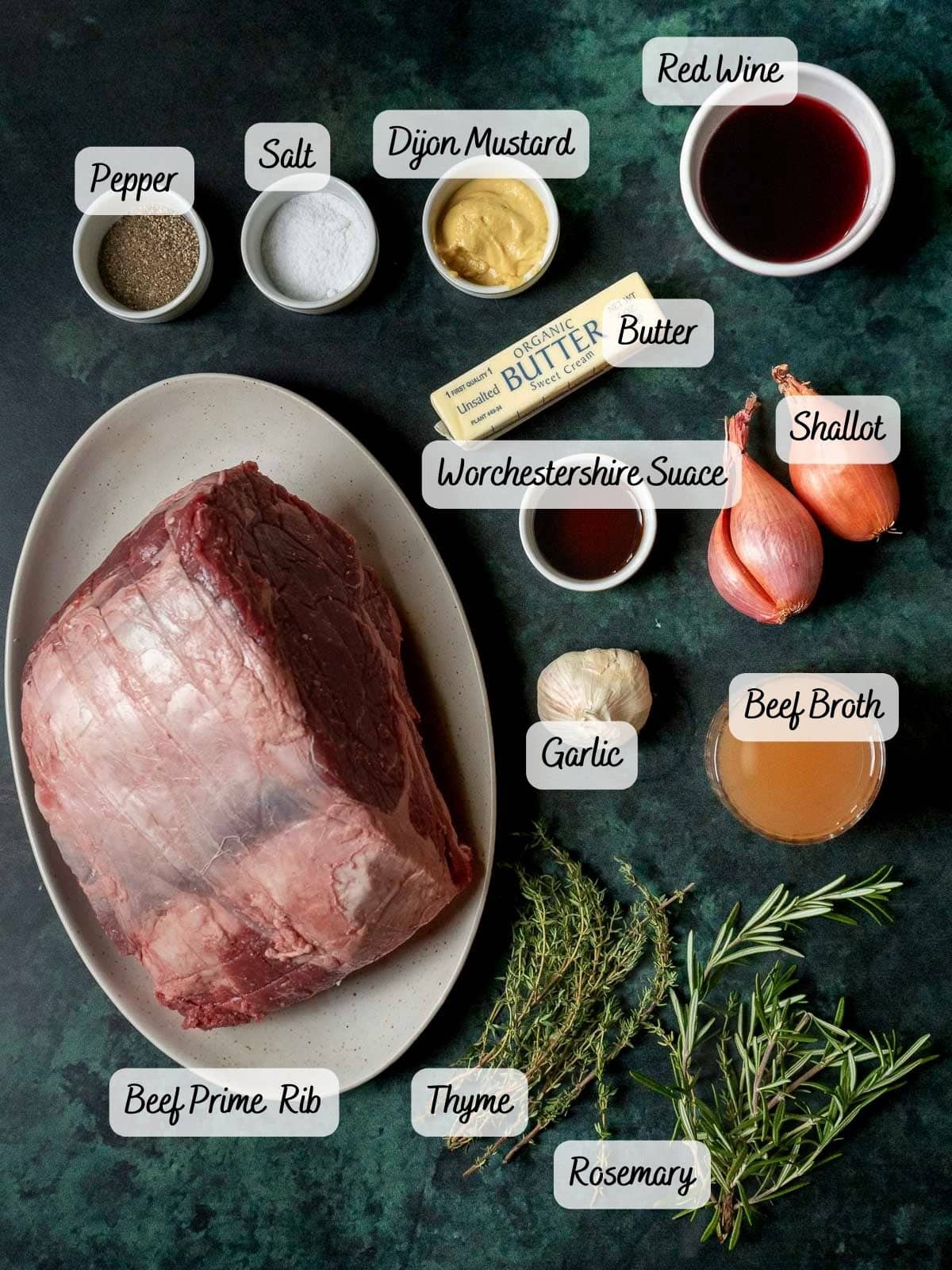 Ingredients for a beef dish: sous vide prime rib, pepper, salt, Dijon mustard, red wine, butter, shallot, Worcestershire sauce, garlic, beef broth, thyme, and rosemary on a dark surface.