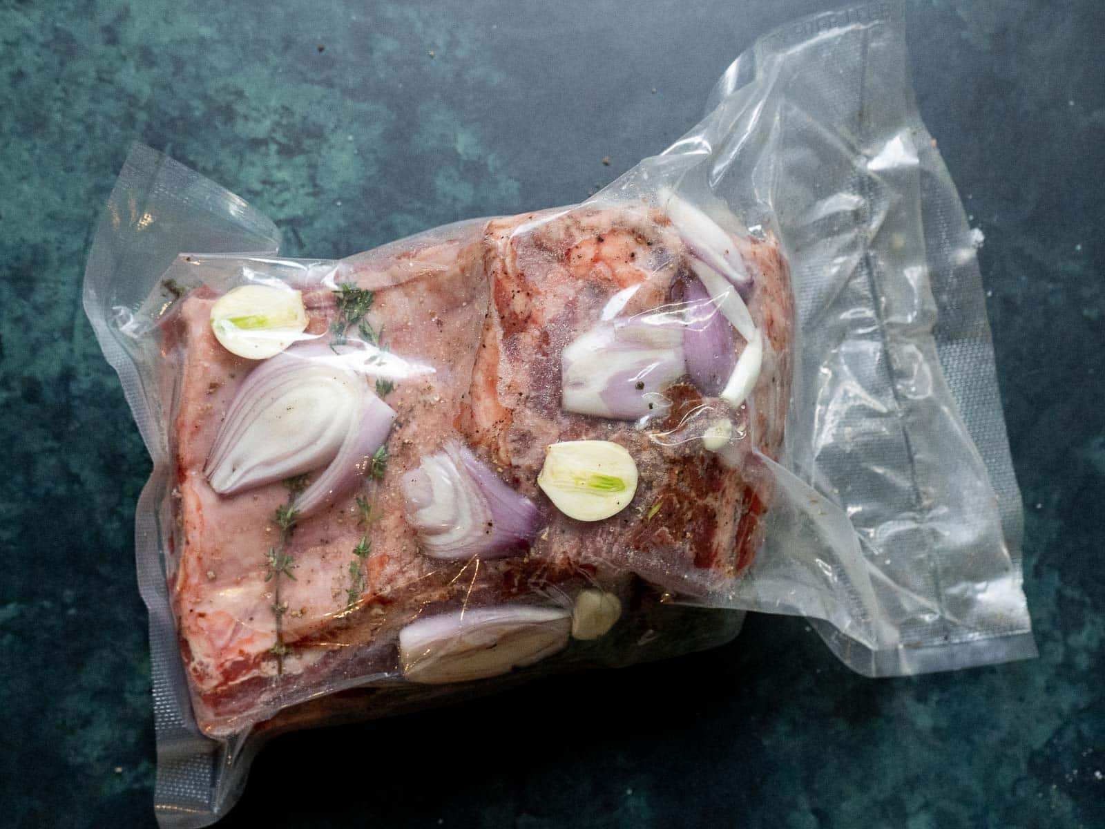 Vacuum-sealed sous vide prime rib with sliced onions, garlic, and herbs rests elegantly on a dark surface.