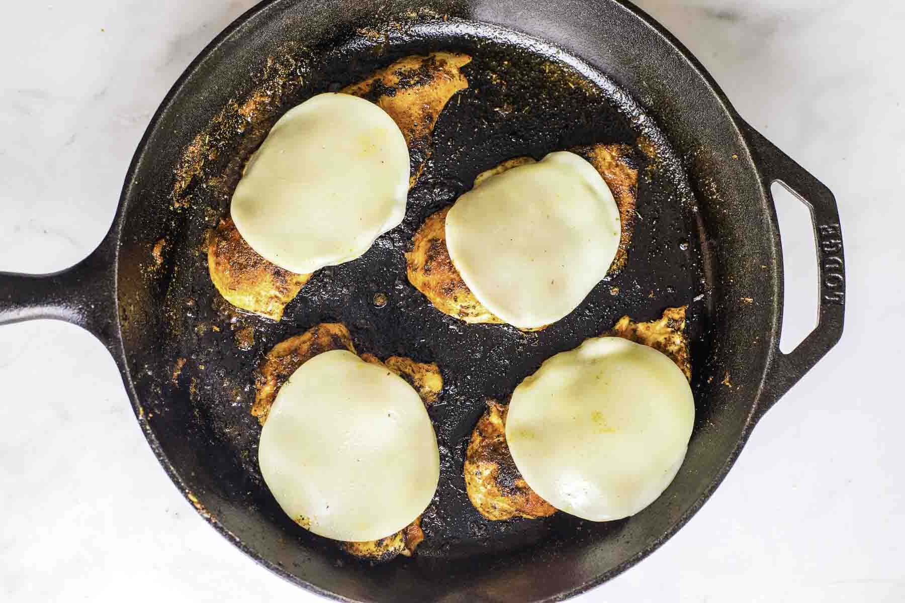 Four chicken breasts with melted cheese slices in a cast iron skillet.