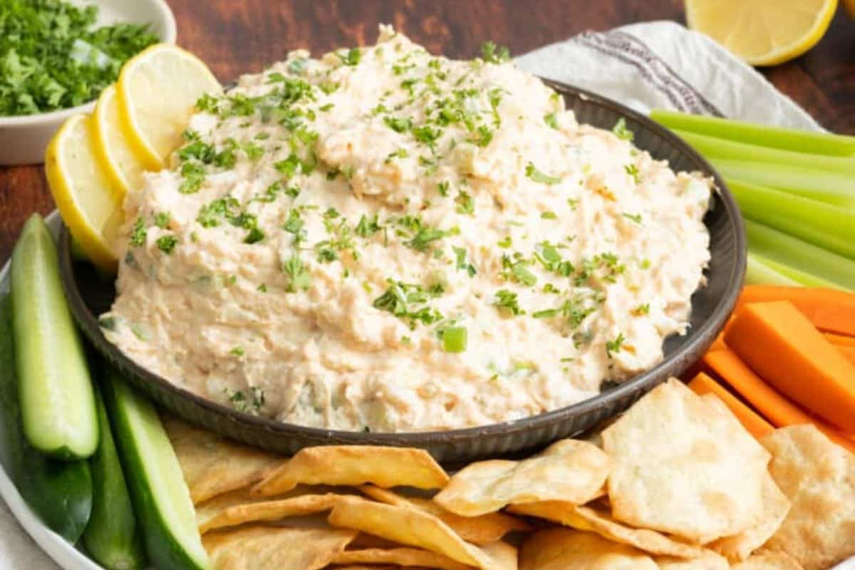 A bowl of creamy dip garnished with parsley, surrounded by lemon wedges, celery sticks, carrot sticks, cucumber slices, and chips on a plate.