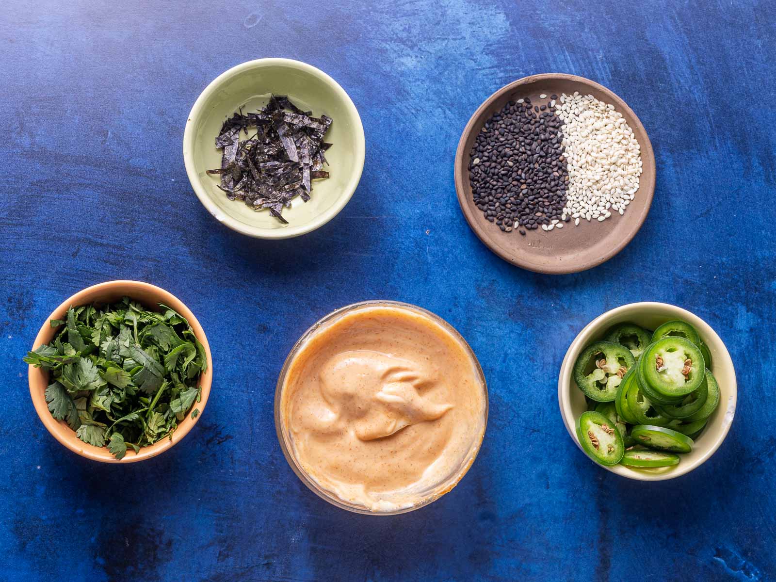 Various small bowls of ingredients on a blue surface, including dried seaweed, black and white sesame seeds, fresh cilantro, creamy sauce, and sliced green jalapeños.