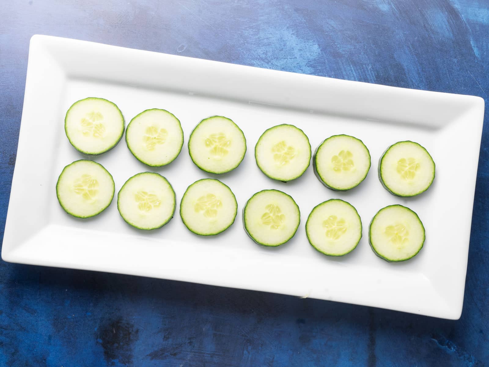 Sliced cucumber arranged in two rows on a rectangular white platter against a blue background.