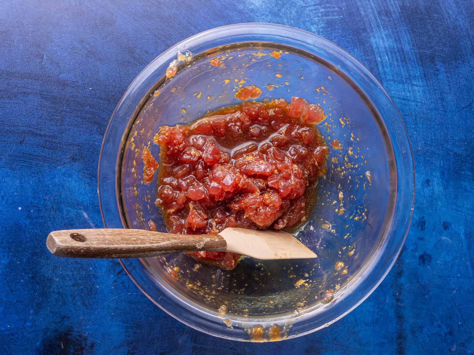 Glass bowl with chopped tomatoes and sauce, mixed with a spatula, on a blue surface.
