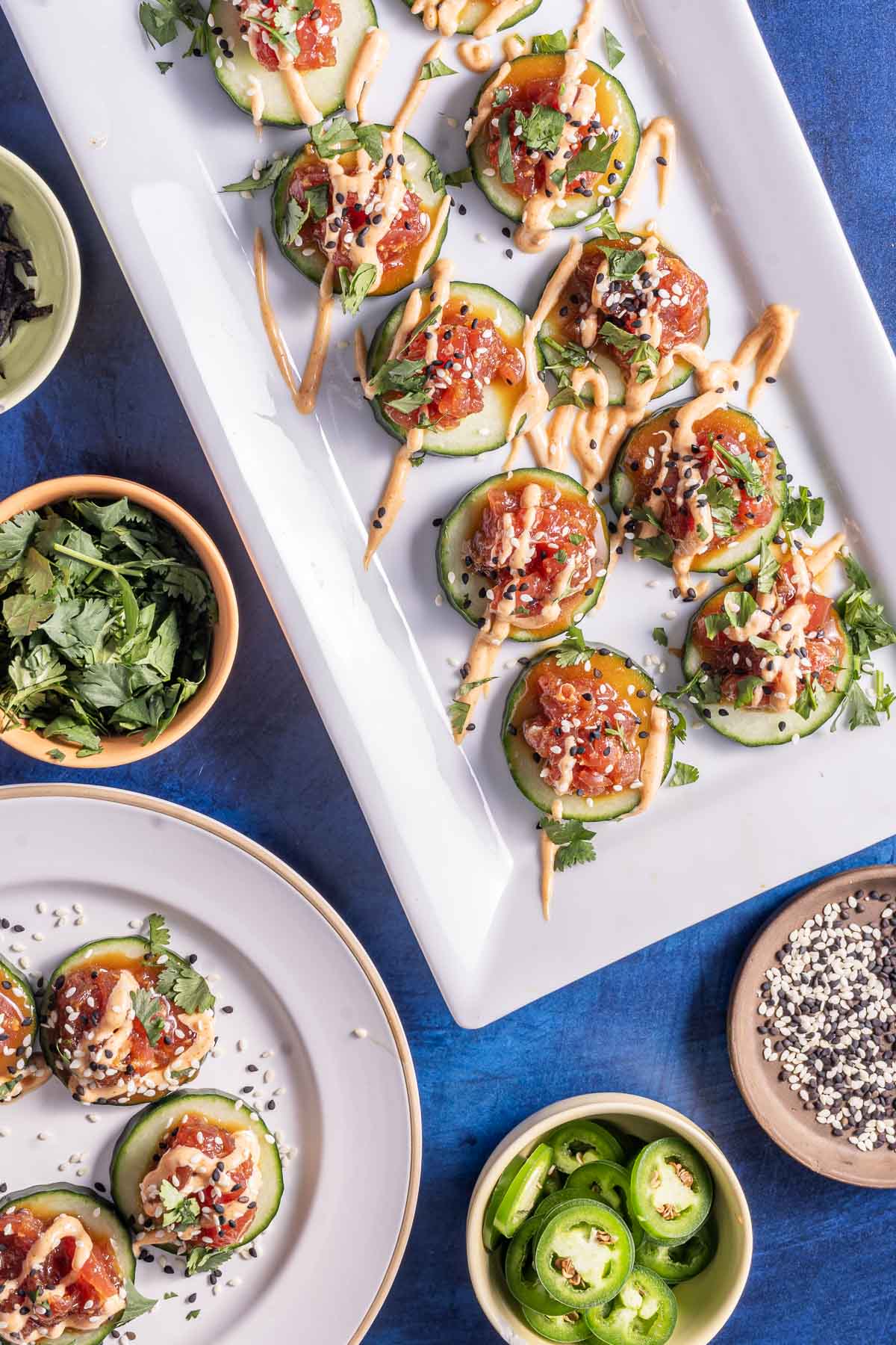 Plates of cucumber sushi topped with diced fish, sesame seeds, and sauce, garnished with cilantro. Nearby bowls contain sliced jalapeños, sesame seeds, and herbs.