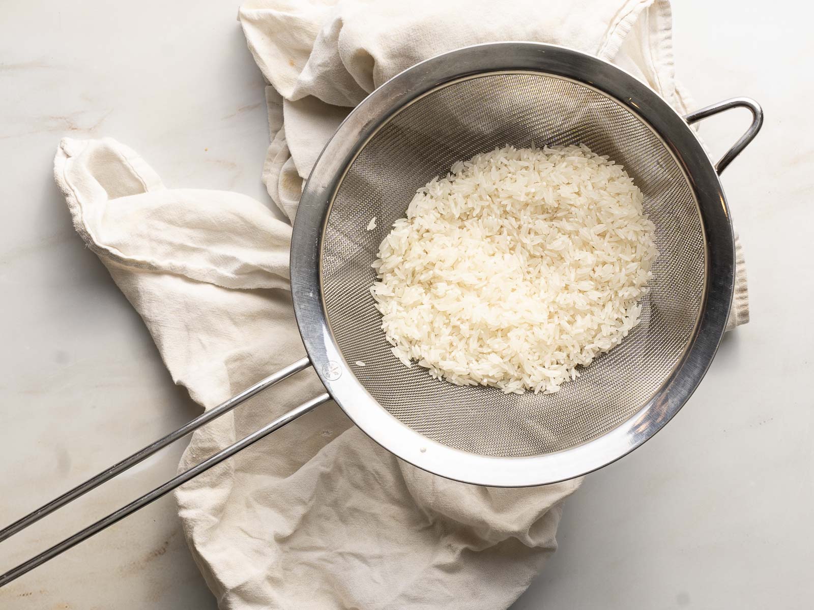 A metal sieve filled with white rice rests on a white cloth on a light surface.