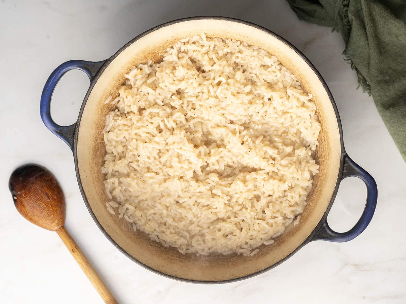Cooked white rice in a blue-handled pot on a light surface, next to a wooden spoon and a green cloth.