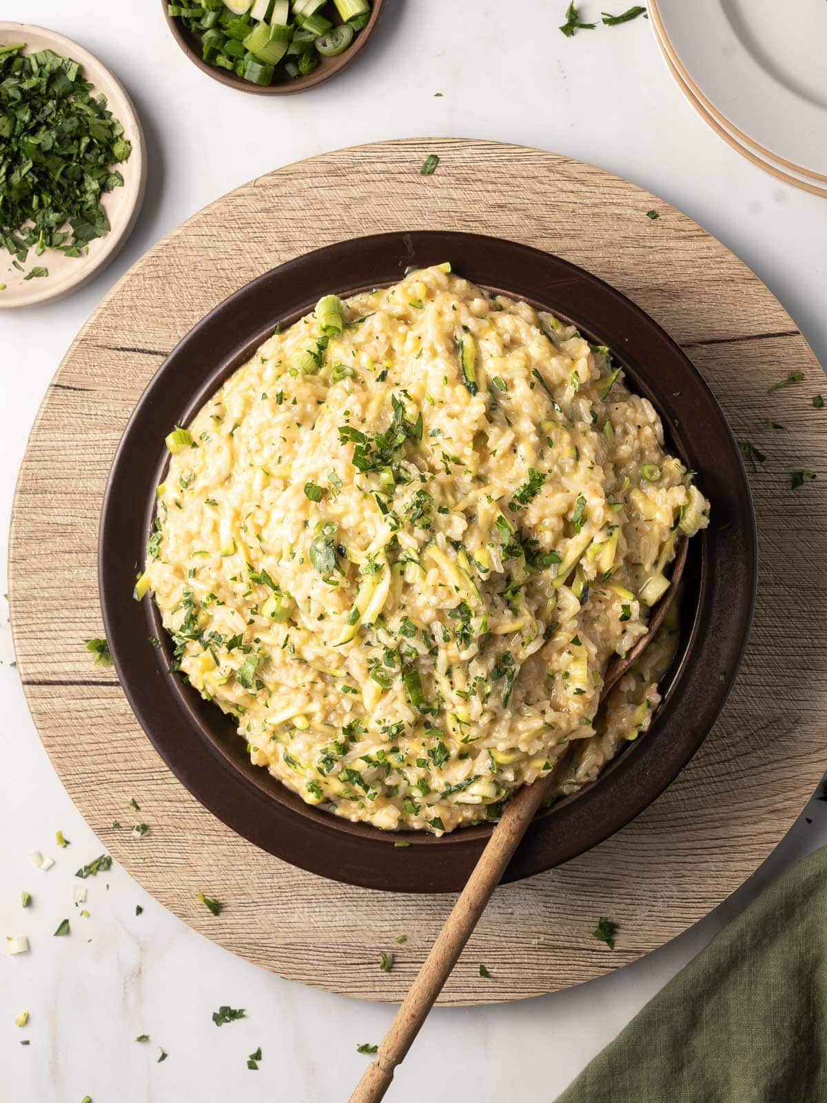 A bowl of creamy coleslaw topped with chopped green herbs, placed on a wooden tray. A wooden spoon rests in the bowl, with a small dish of herbs and stacked plates nearby.