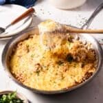 A skillet with a cheesy rice dish topped with melted cheese and herbs, being served with a spoon. Plates and a napkin are set nearby.