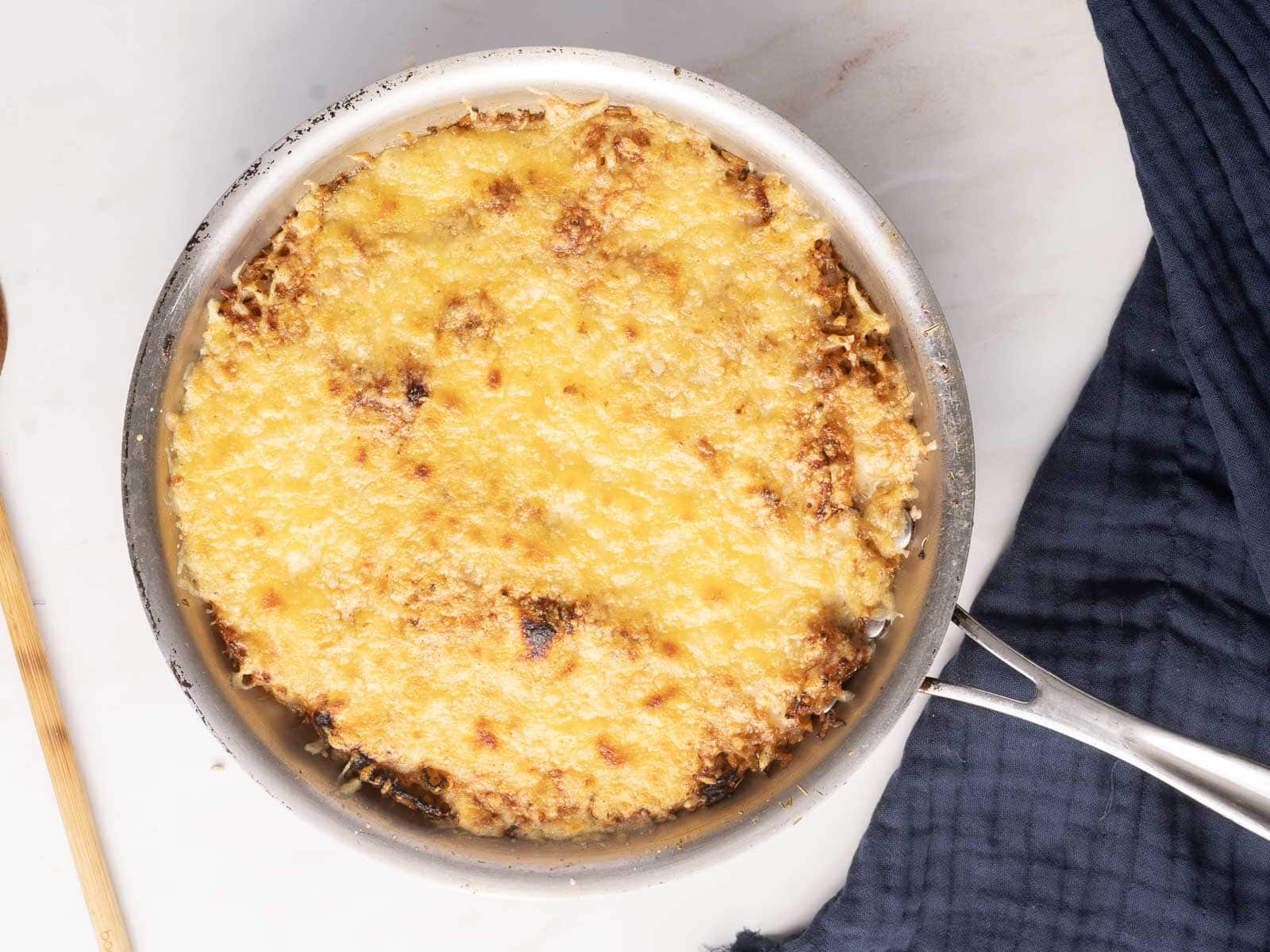 A skillet with a baked, golden-brown cheese topping, likely a gratin or casserole, on a marble surface next to a blue cloth.