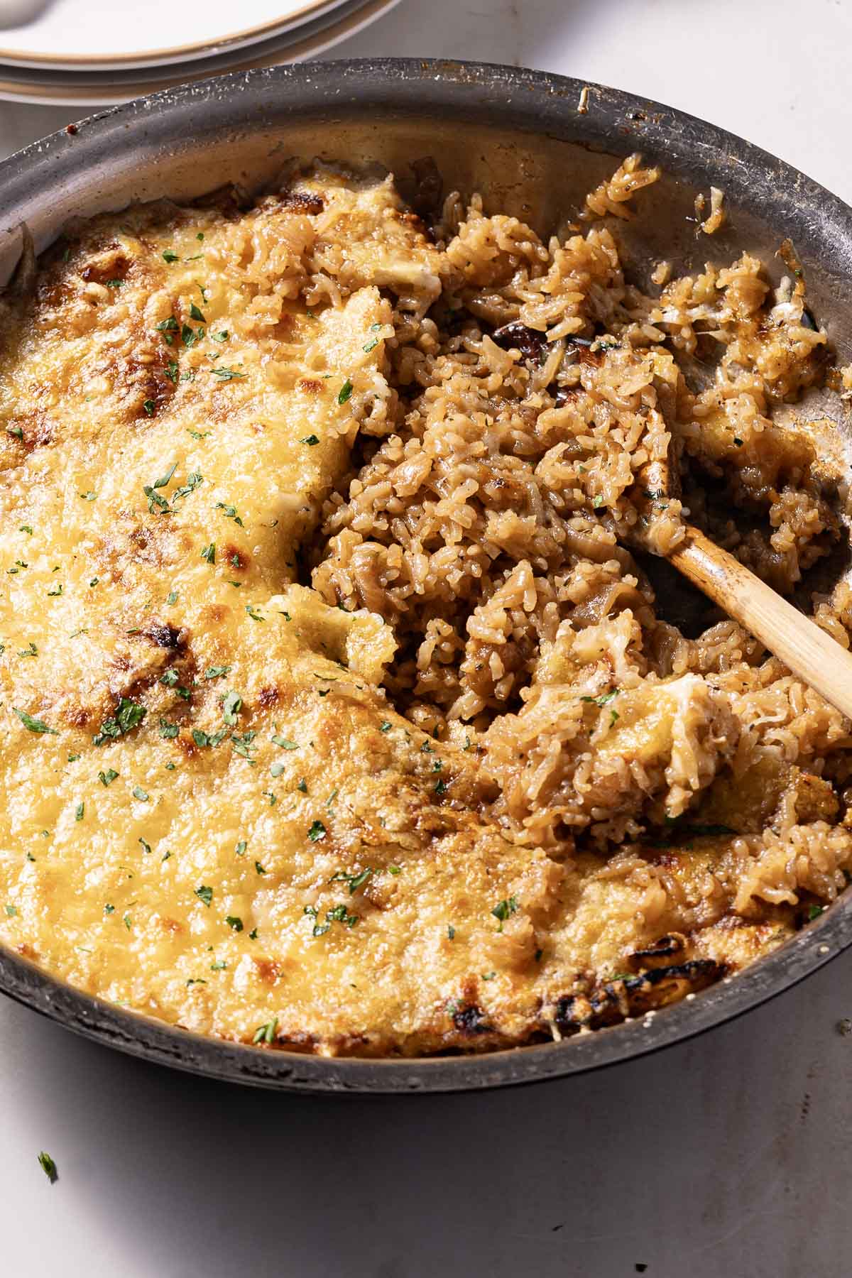 A skillet filled with cheesy baked rice, garnished with herbs, and a wooden spoon stirring through it.