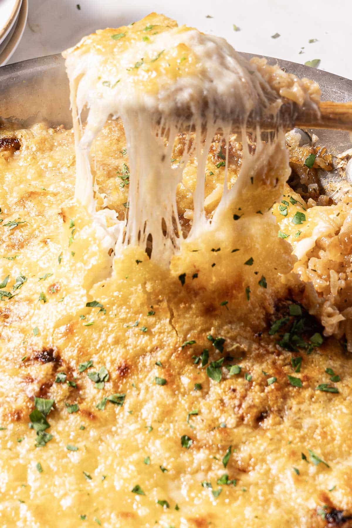 A cheesy, baked dish garnished with chopped parsley, with a portion being lifted by a wooden spoon, showing melted cheese stretching.