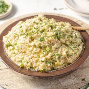 A brown plate of zucchini risotto garnished with chopped herbs on a light wooden surface.