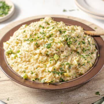 A brown plate of zucchini risotto garnished with chopped herbs on a light wooden surface.