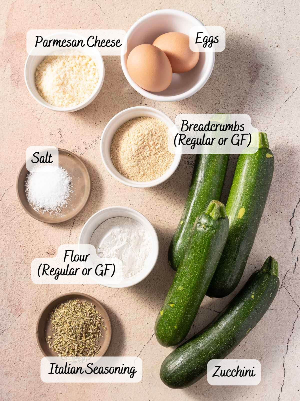Ingredients for a dish: zucchini, eggs, breadcrumbs, flour, parmesan cheese, Italian seasoning, and salt arranged on a light surface with labels.