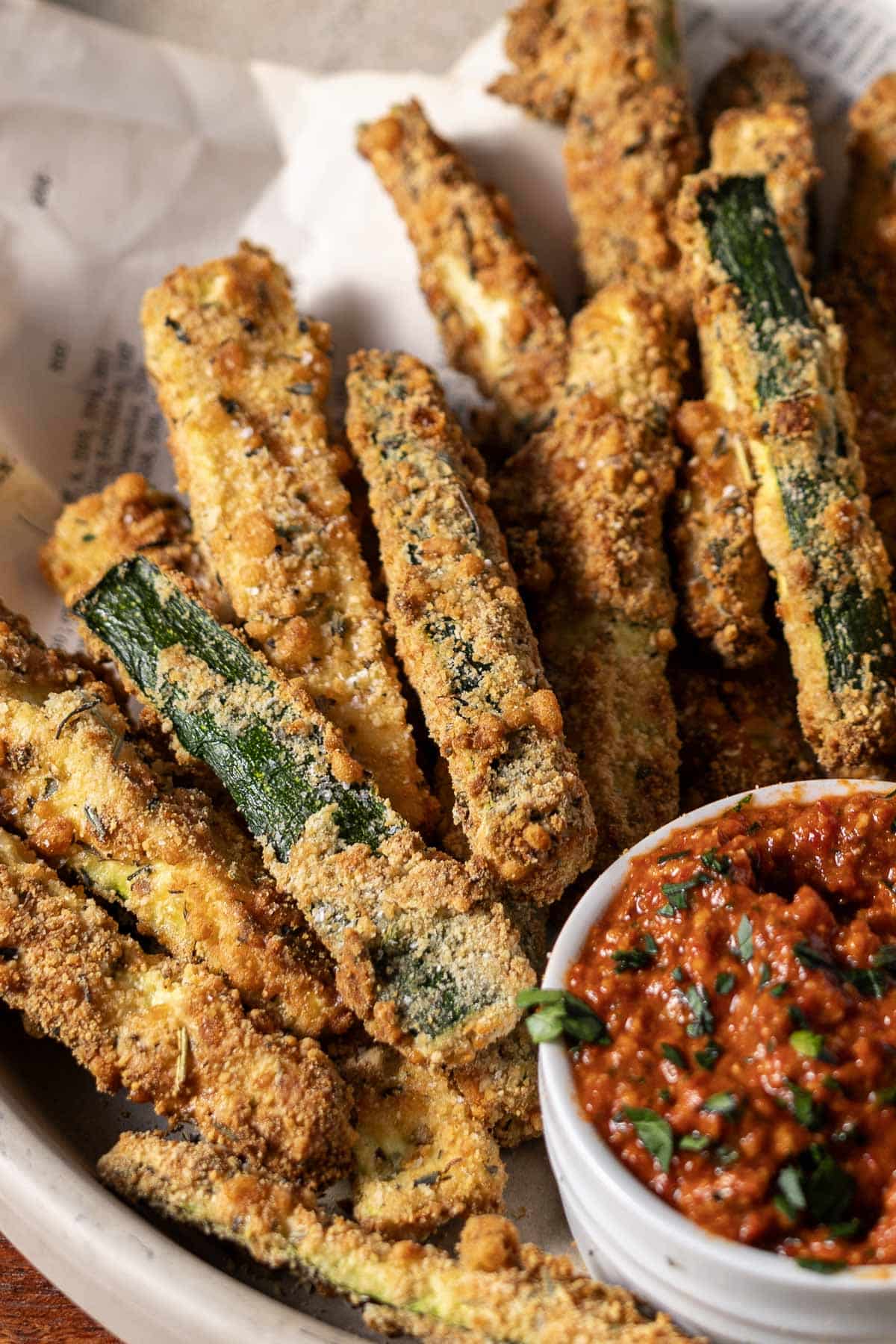A plate of breaded zucchini fries next to a small bowl of red dipping sauce garnished with herbs.